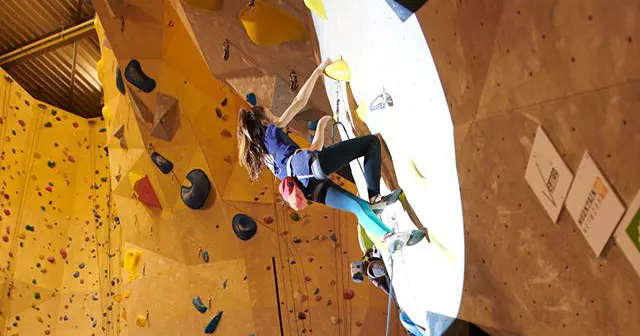Iemand beklimt een indoor-klimwand en reikt met één hand naar een gele greep, terwijl hij vastzit in een klimgordel. De wand is voorzien van grepen in verschillende kleuren en schuine vlakken. Felle verlichting zet de klimmer in het licht.
